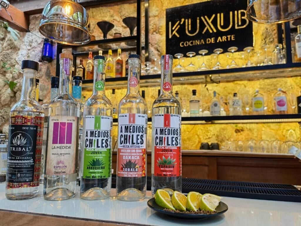 Un comptoir de bar avec différentes bouteilles d’alcool, une enseigne indiquant « K’UXUB Cocina de Arte », et une assiette de tranches de citron vert.