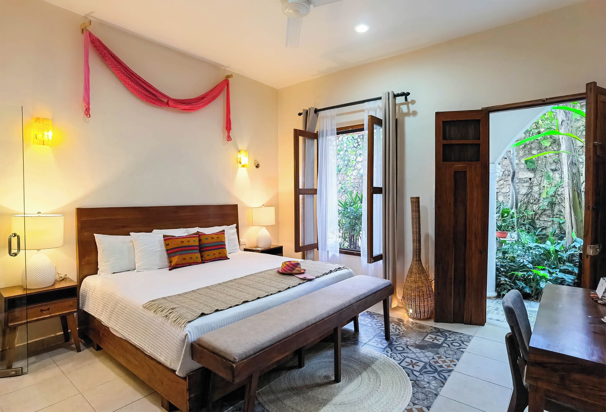 A hotel room with a bed, bedside lamps, a bench, a dining table, and a view of greenery outside.