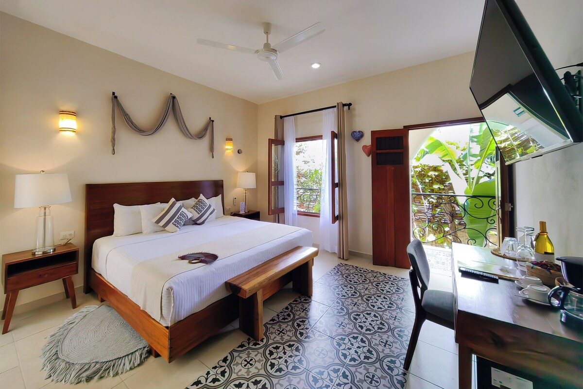 A hotel room with a bed, bedside lamps, a desk with a chair, a ceiling fan, and a view of greenery outside.