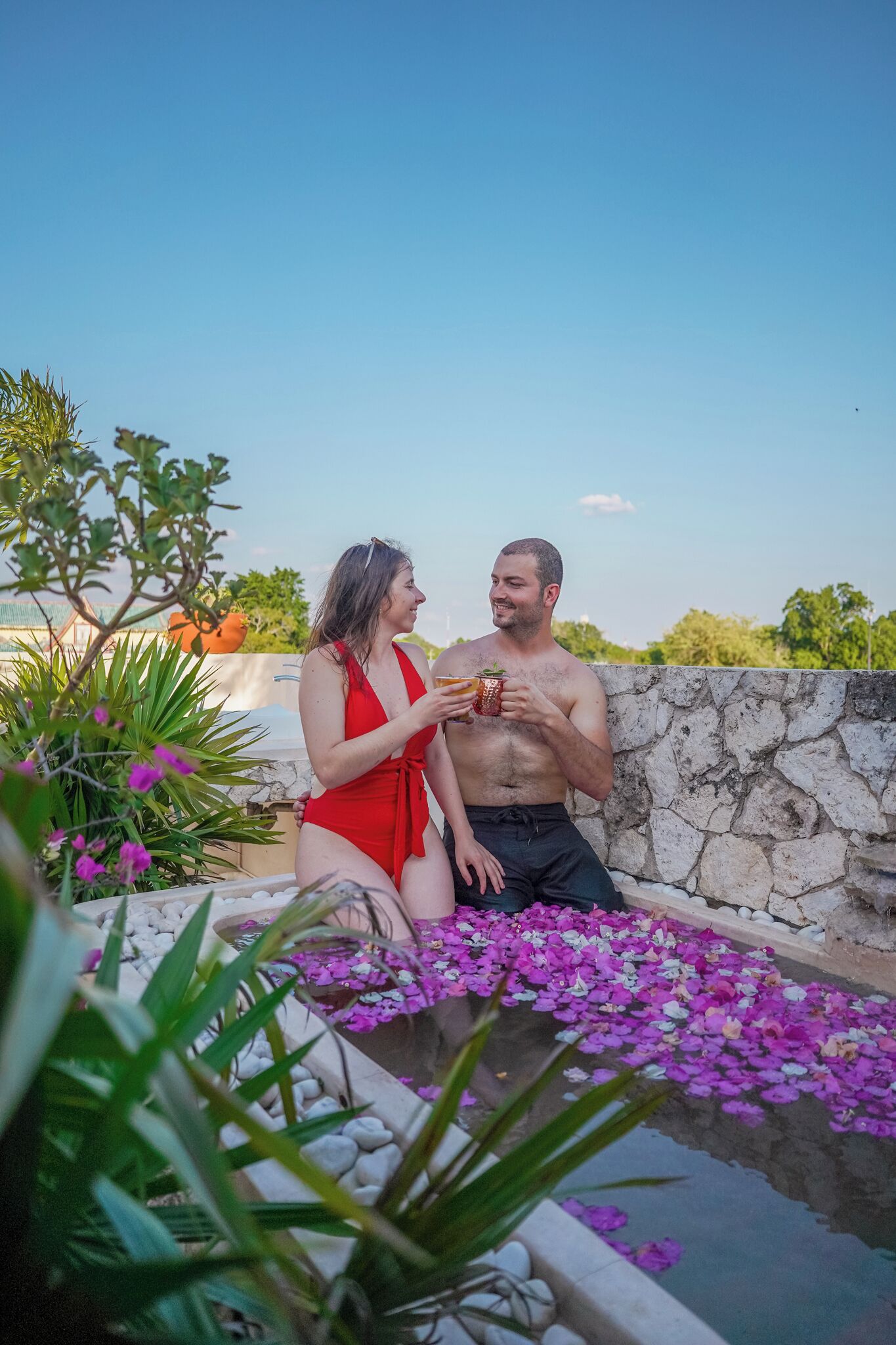 Una parella asseguda a la piscina, amb nenúfars, envoltada de verdura i una paret de pedra.