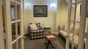 A cozy room with a checkered armchair, a small table with a candle, and a sofa with patterned cushions. A framed painting of a sailboat hangs on the wall.