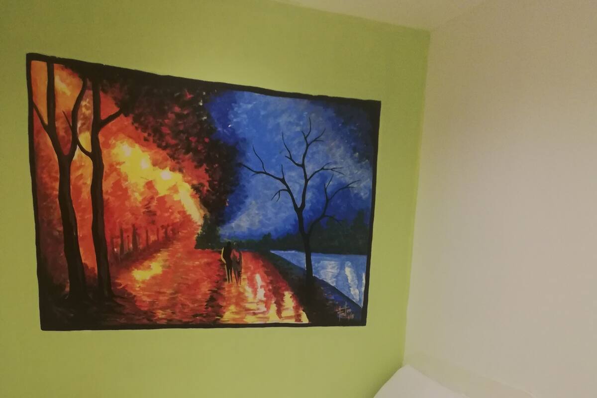 A room with a bed against a wall, a green wall, and a painting of a forest scene.