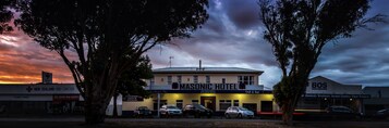 Hotel Masonic com uma placa, um estacionamento com vários carros e edifícios adjacentes, incluindo um da Cruz Vermelha e da BOS Cleaning Supplies.
