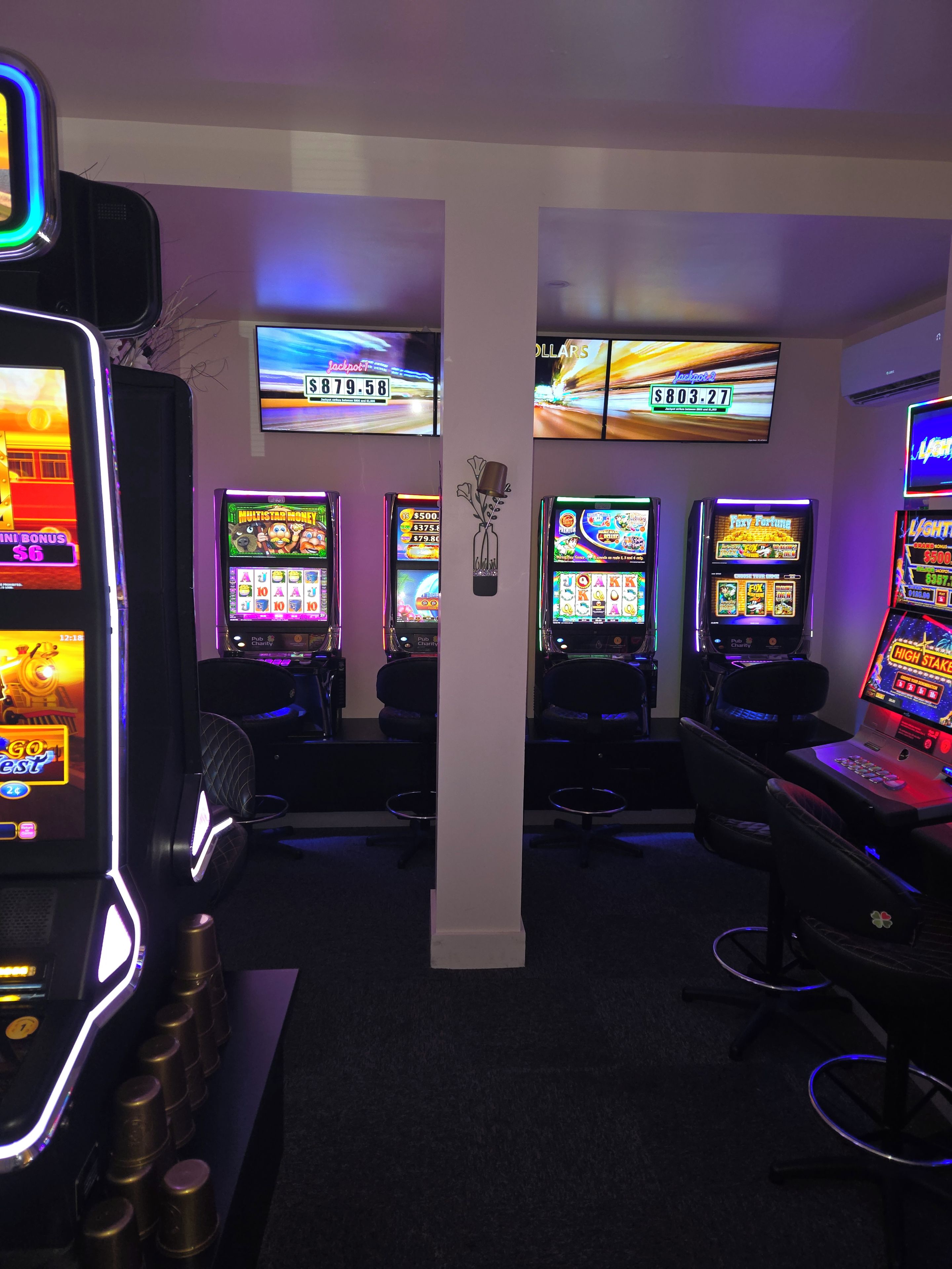 A row of slot machines with digital displays, a pillar with a floral decoration, and a wall-mounted television.