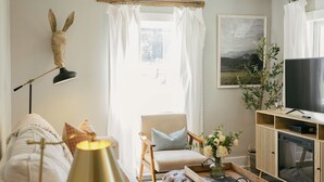 Ett mysigt vardagsrum med en soffa, en pall och ett soffbord. Där finns också en lampa, ett brickbord med böcker och en tv.