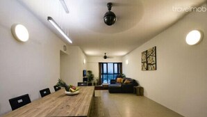 A spacious living room with a wooden dining table, black chairs, a sofa with cushions, a ceiling fan, and a large window providing natural light.