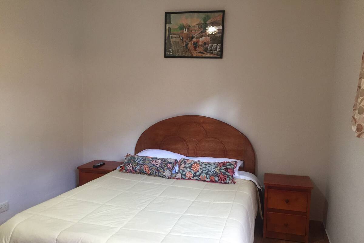 Ein ordentlich bezogenes Bett mit weißer Daunendecke und einem blumigen Kopfkissen, ein Holzkopfbrett, ein Nachttisch mit Fernbedienung und ein an der Wand hängendes Bild von einer Straßenszene.