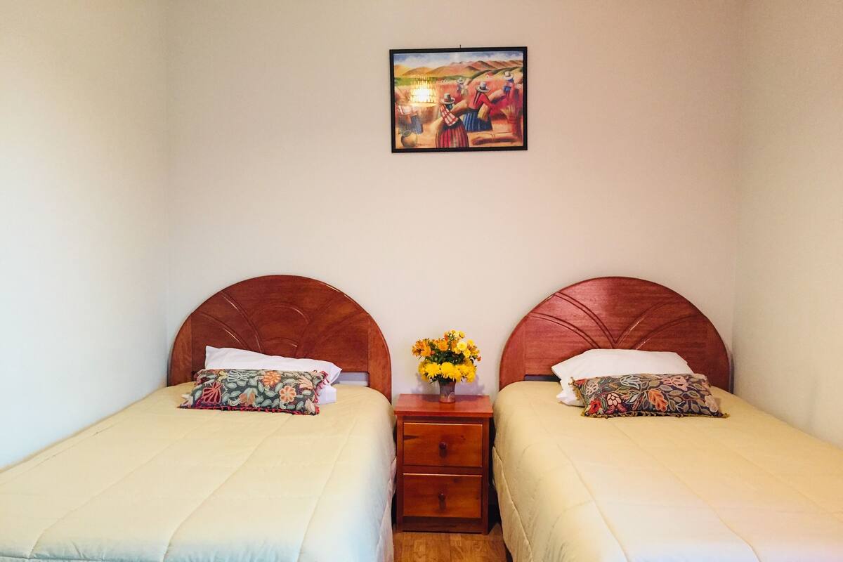 Ein Doppelzimmer mit zwei Betten, jeweils mit einem Holz-Kopfteil, einem Nachttisch mit Blumenvase und einem Bild an der Wand.