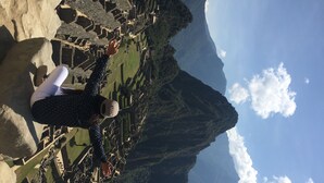 A person standing on a stone platform overlooking a terraced landscape with ancient ruins and a mountain in the background.