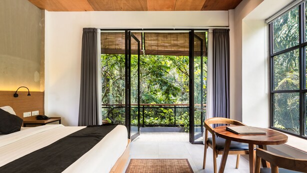 Une chambre d’hôtel moderne, dotée d’un grand lit, d’un bureau en bois et offrant une vue sur une végétation luxuriante grâce à une porte-fenêtre coulissante.
