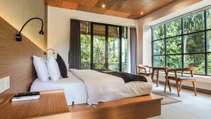 Une chambre d’hôtel moderne avec un plafond en bois, un grand lit avec une literie blanche, une table de chevet avec une télécommande et une vue sur une végétation luxuriante, visible à travers de grandes fenêtres.