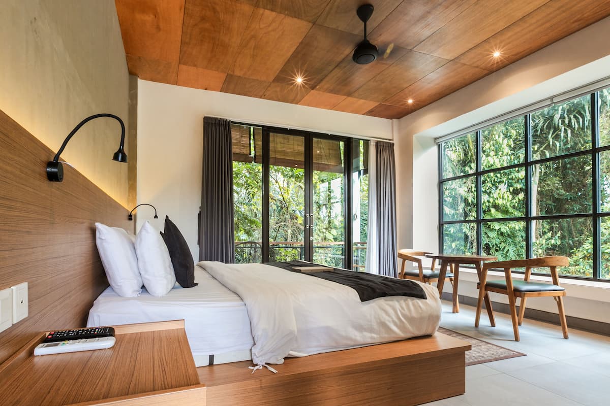 Une chambre d’hôtel moderne avec un plafond en bois, un grand lit avec une literie blanche, une table de chevet avec une télécommande et une vue sur une végétation luxuriante, visible à travers de grandes fenêtres.