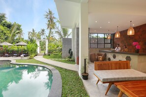 A modern outdoor pool area with a curved edge, surrounded by lush greenery and palm trees. There is a wooden bench and a table near the pool, and a stone wall with a window and a hanging light fixture. - The Nani (Ubud)