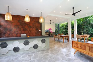 A reception area with a stone wall, hexagonal tiles, and a wooden reception desk. There are pendant lights, ceiling fans, and a view of greenery outside. - The Nani (Ubud)