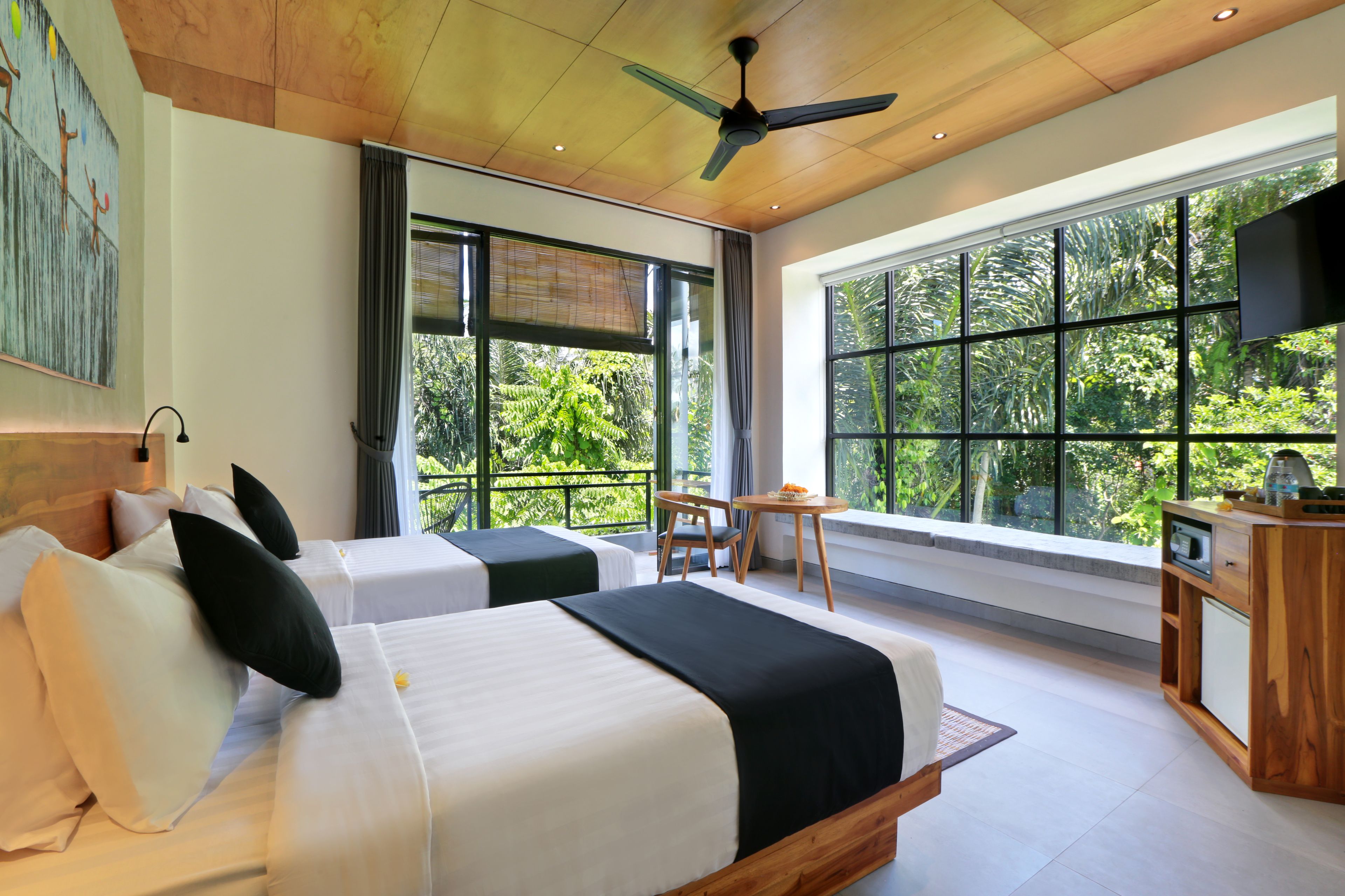 A modern hotel room with two beds, a ceiling fan, large windows, a small table, and a TV.