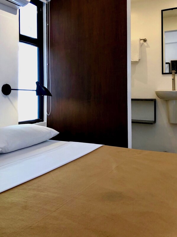 A hotel room with a bed, a window with blinds, a wooden door, a sink, and a mirror. - Gomez House (Makati)