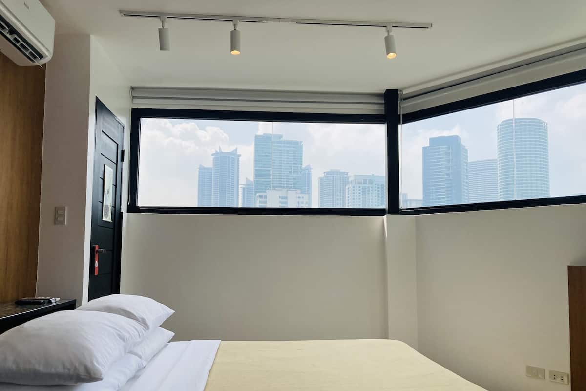 A hotel room with a bed, a window with a city view, an air conditioning unit, and a bedside table.