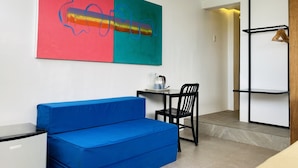 A room with a blue sofa, a small desk with a chair, a large abstract painting, and a wooden shelf unit. - Gomez House (Makati)