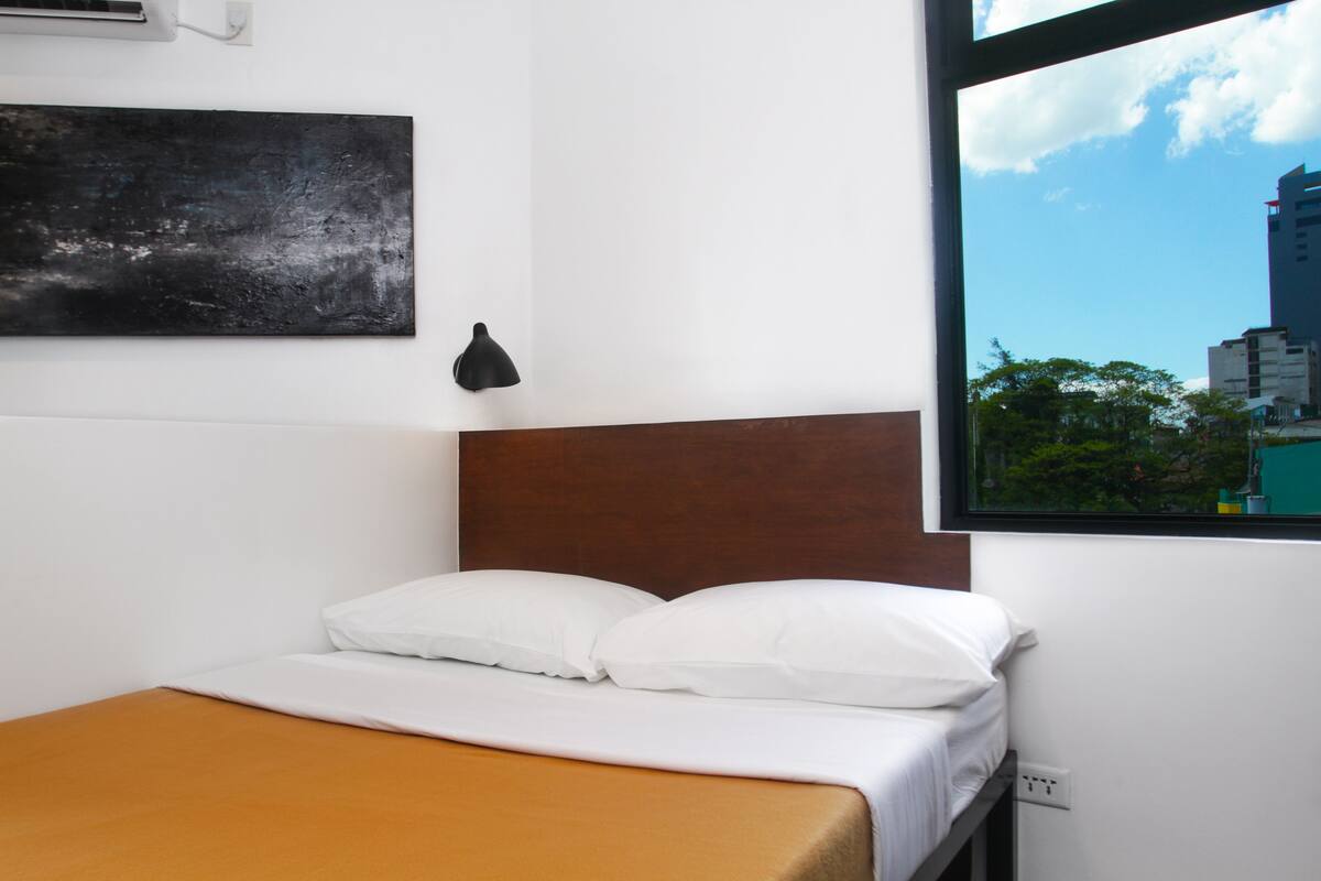 A hotel room with a bed, a window with a city view, a wall-mounted air conditioner, and a black artwork piece.