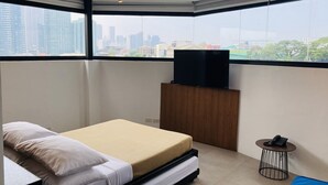 A bedroom with a bed, a TV on a wooden stand, a small round table with a phone, and a city view through large windows. - Gomez House (Makati)