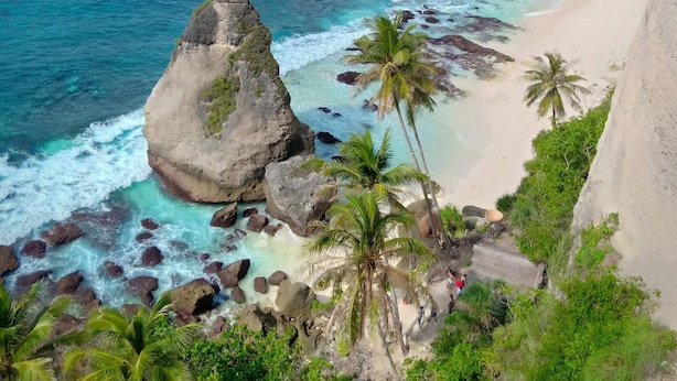 Un paisaje costero con acantilados rocosos, aguas turquesas cristalinas y una playa arenosa con palmeras.