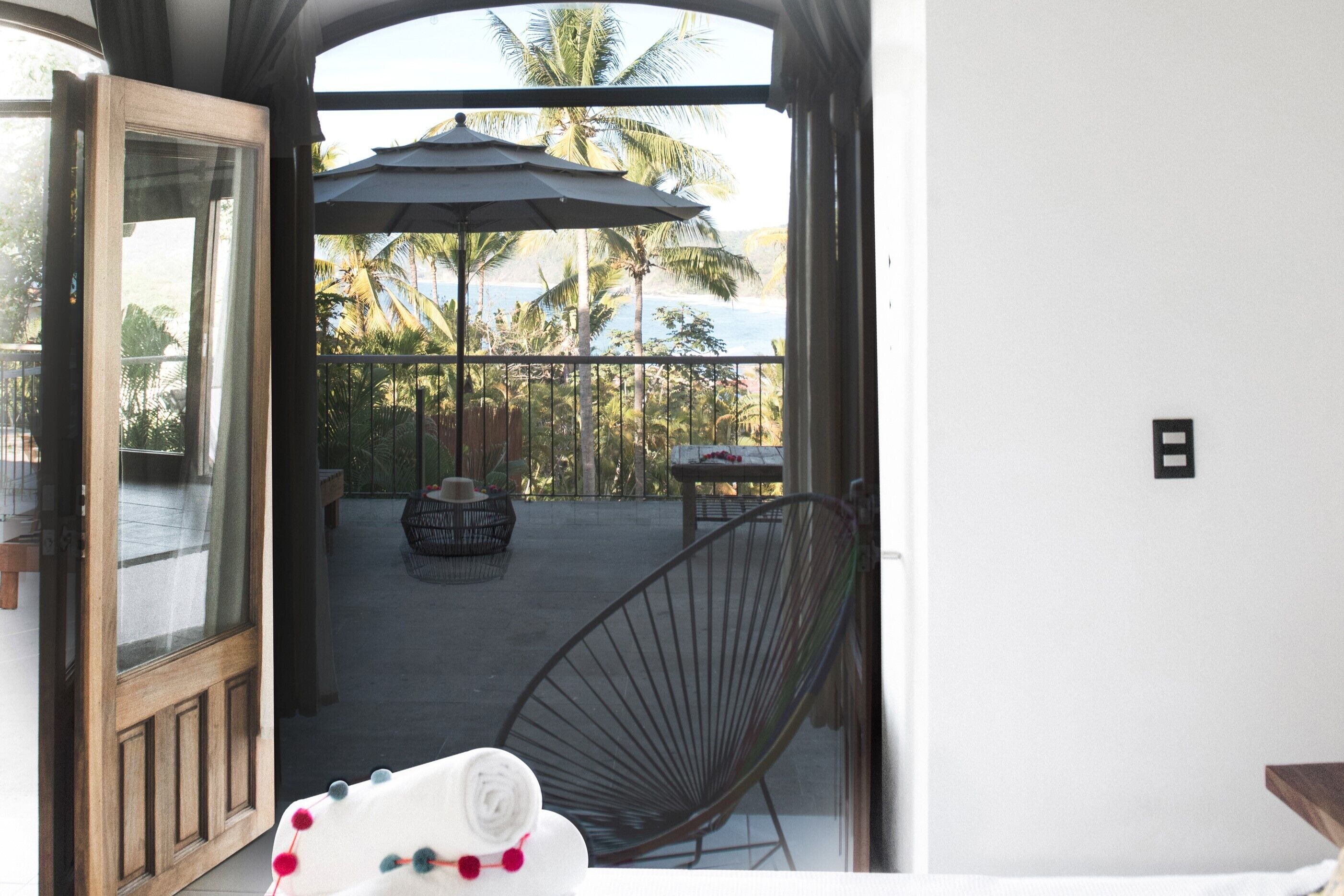 A balcony with a view of palm trees and the sea, a chair, and a table with an umbrella.