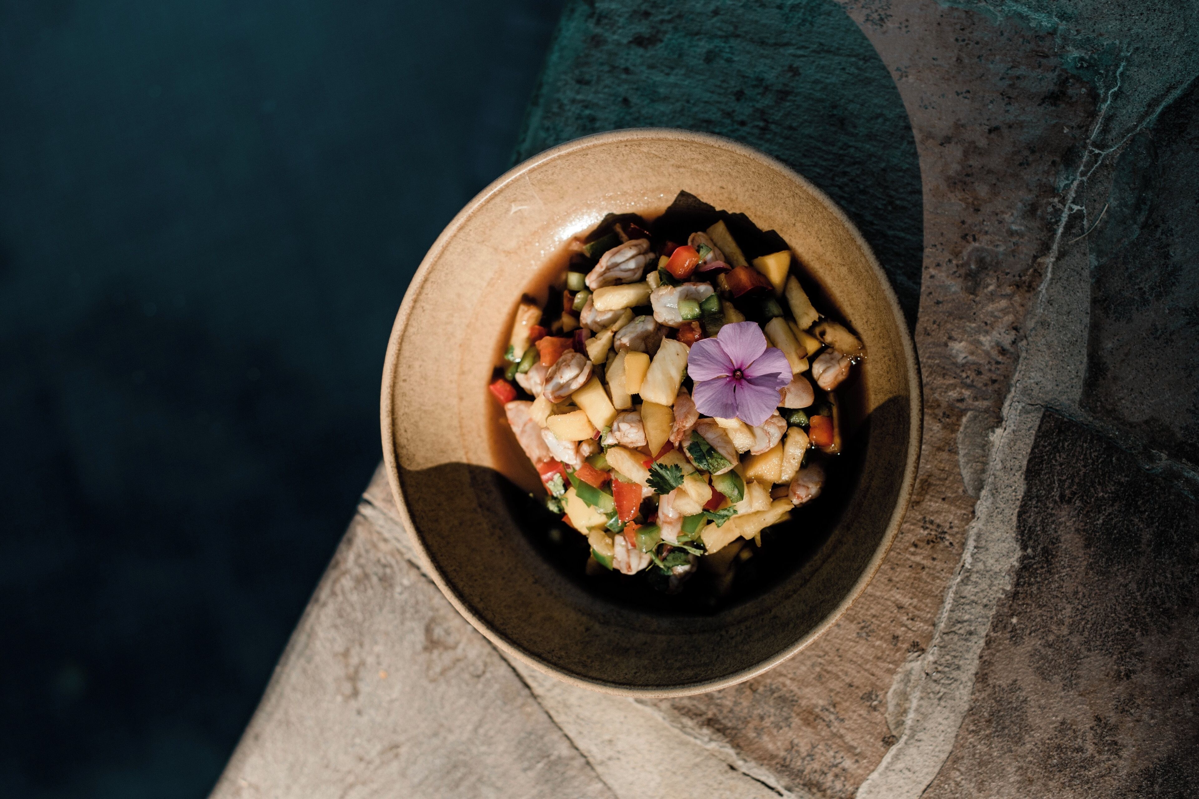 A bowl of fruit salad with a purple flower on a stone surface.