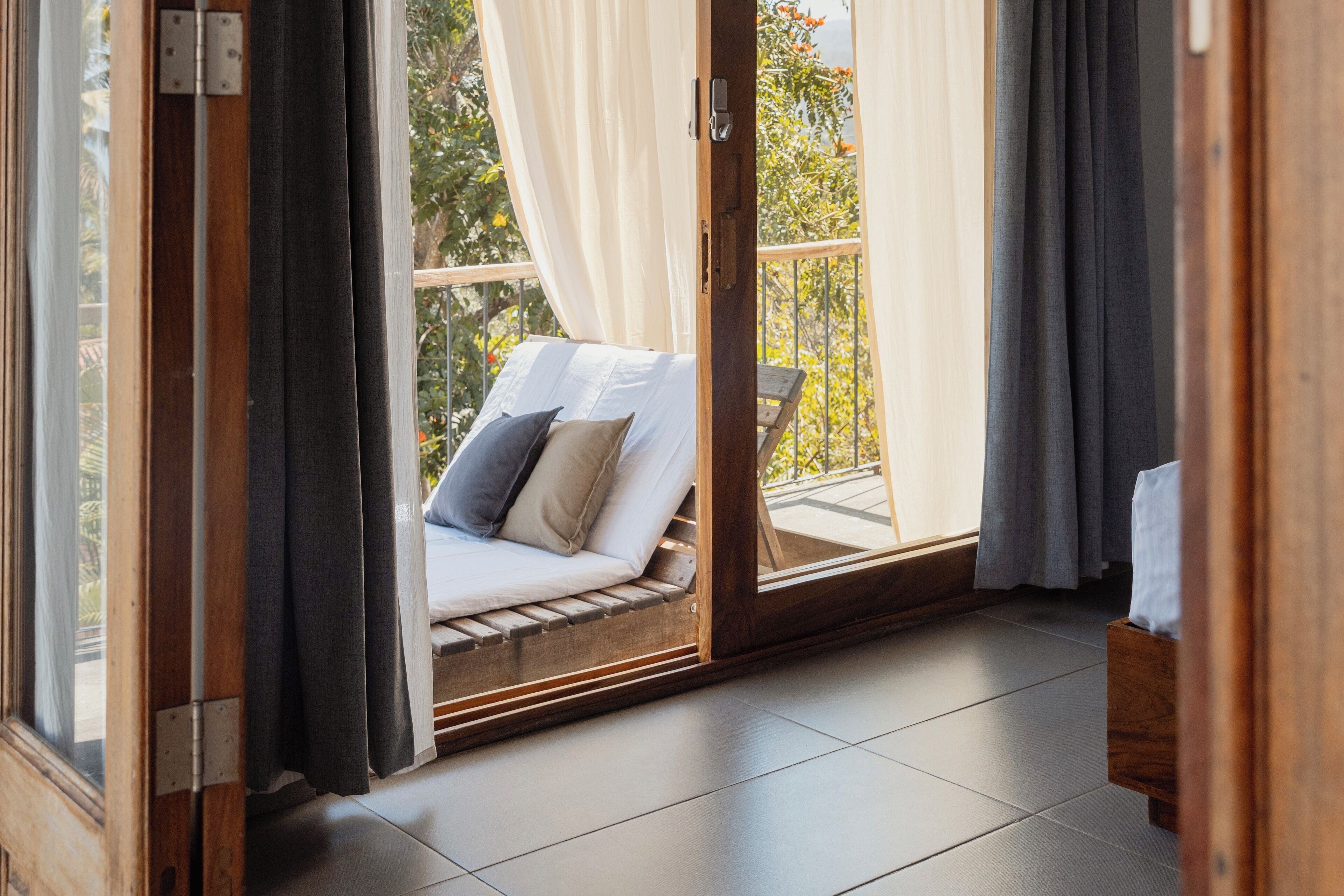 A balcony with a cushioned seating area, a wooden floor, and a view of greenery.