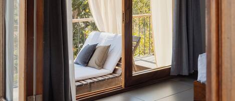 A balcony with a cushioned seating area, a wooden floor, and a view of greenery.