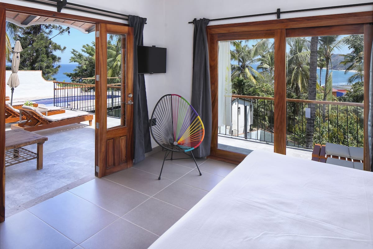 A hotel room with a bed, a colorful chair, a TV, and a view of the ocean and palm trees.