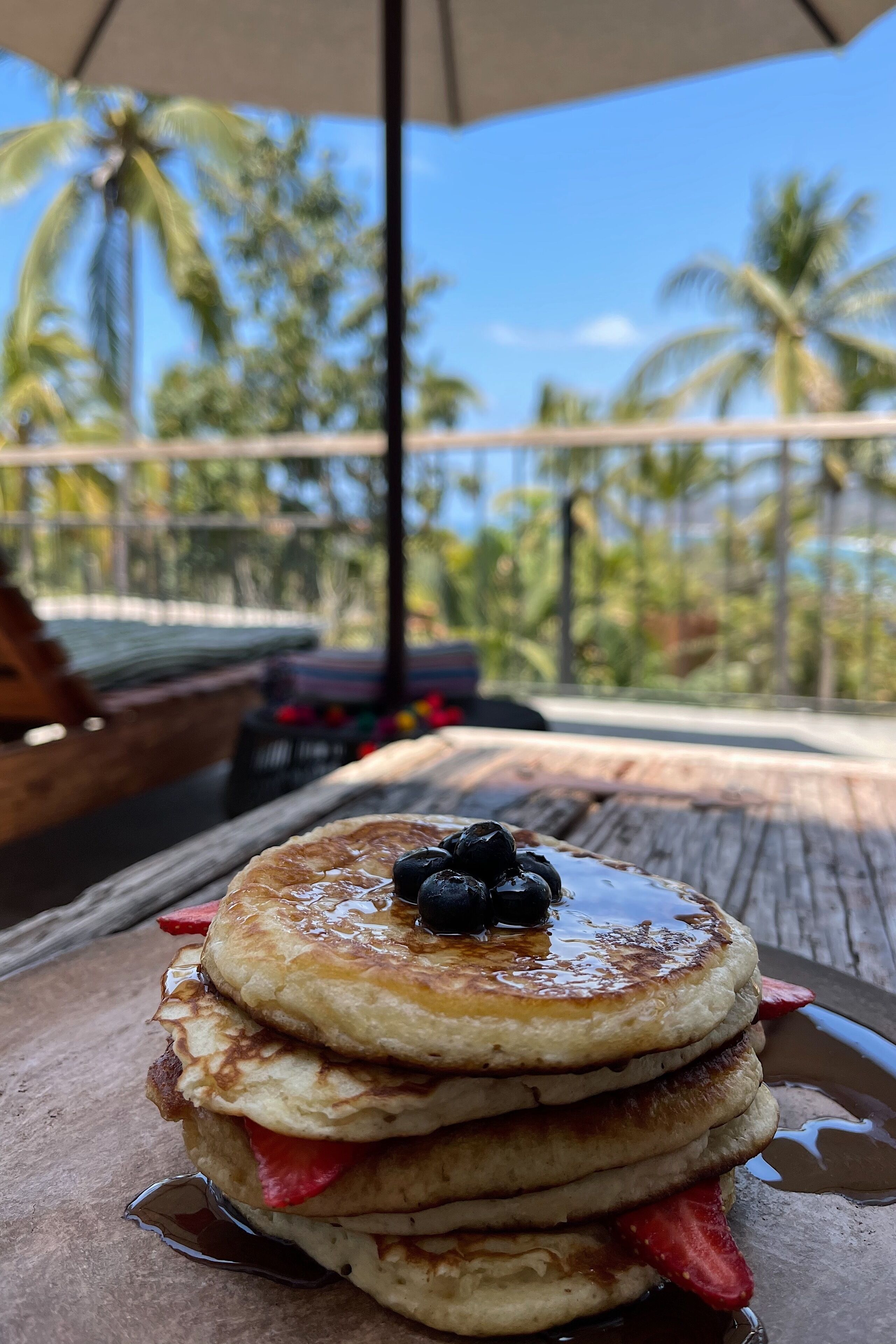 Una pila de tortitas con almíbar y fresas sobre una mesa de madera bajo un paraguas.