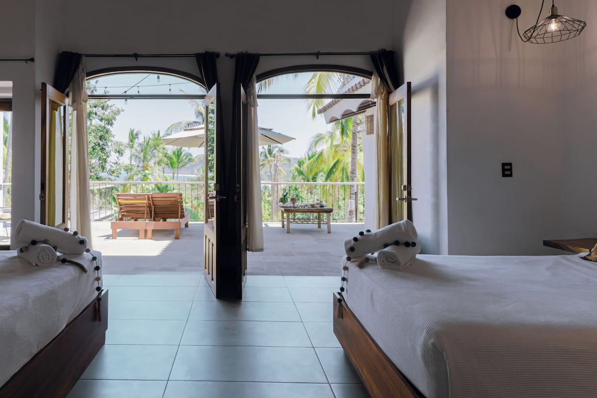 A hotel room with two beds, a balcony with a table and chairs, and a view of palm trees and mountains.