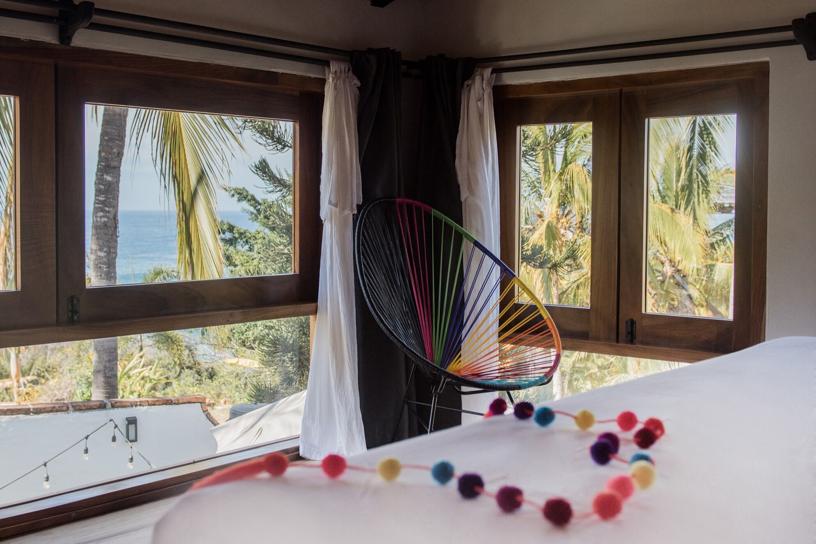 Una habitación con cama, una silla colgante de colores y ventanas amplias con cortinas.