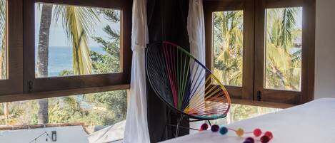 Una habitación con cama, una silla colgante de colores y ventanas amplias con cortinas.