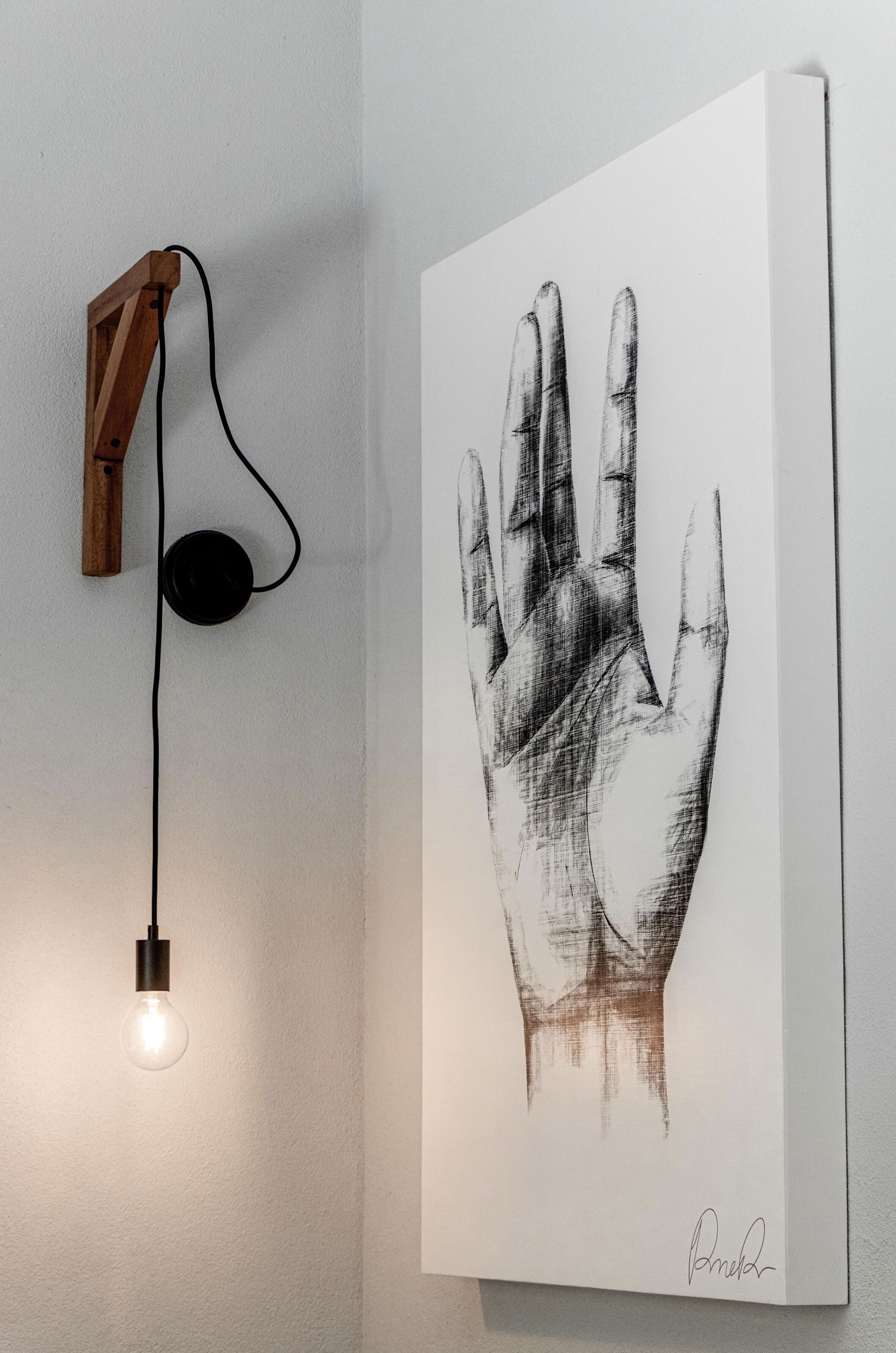 Una lámpara de pared con base de madera y cable negro, junto a una obra de arte con una huella de mano blanco y negro firmada por el artista.