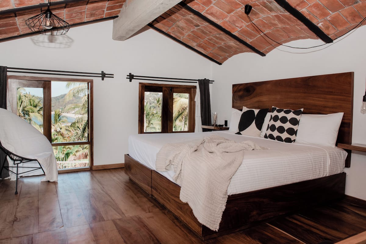 A bedroom with a wooden bed, a chair, and a view of palm trees through large windows.