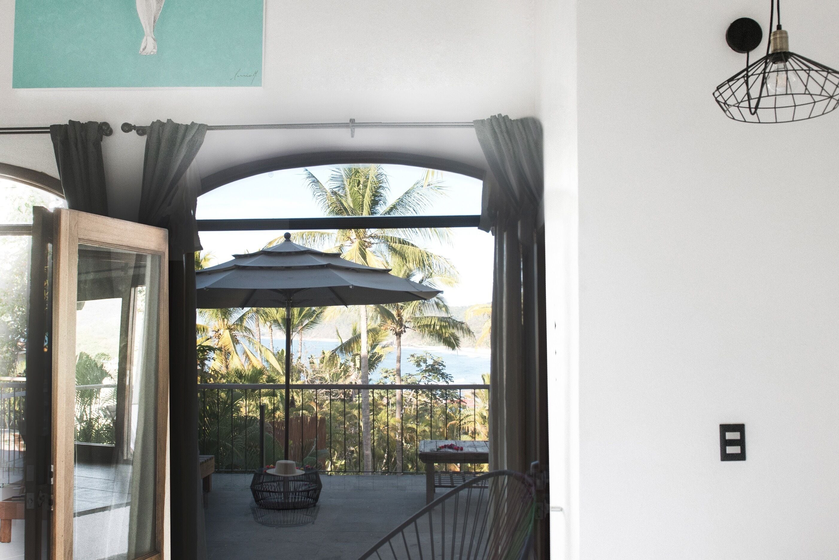 A balcony with a view of palm trees and the ocean, a table with a chair, and a hanging light fixture.