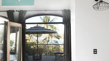 A balcony with a view of palm trees and the ocean, a table with a chair, and a hanging light fixture.