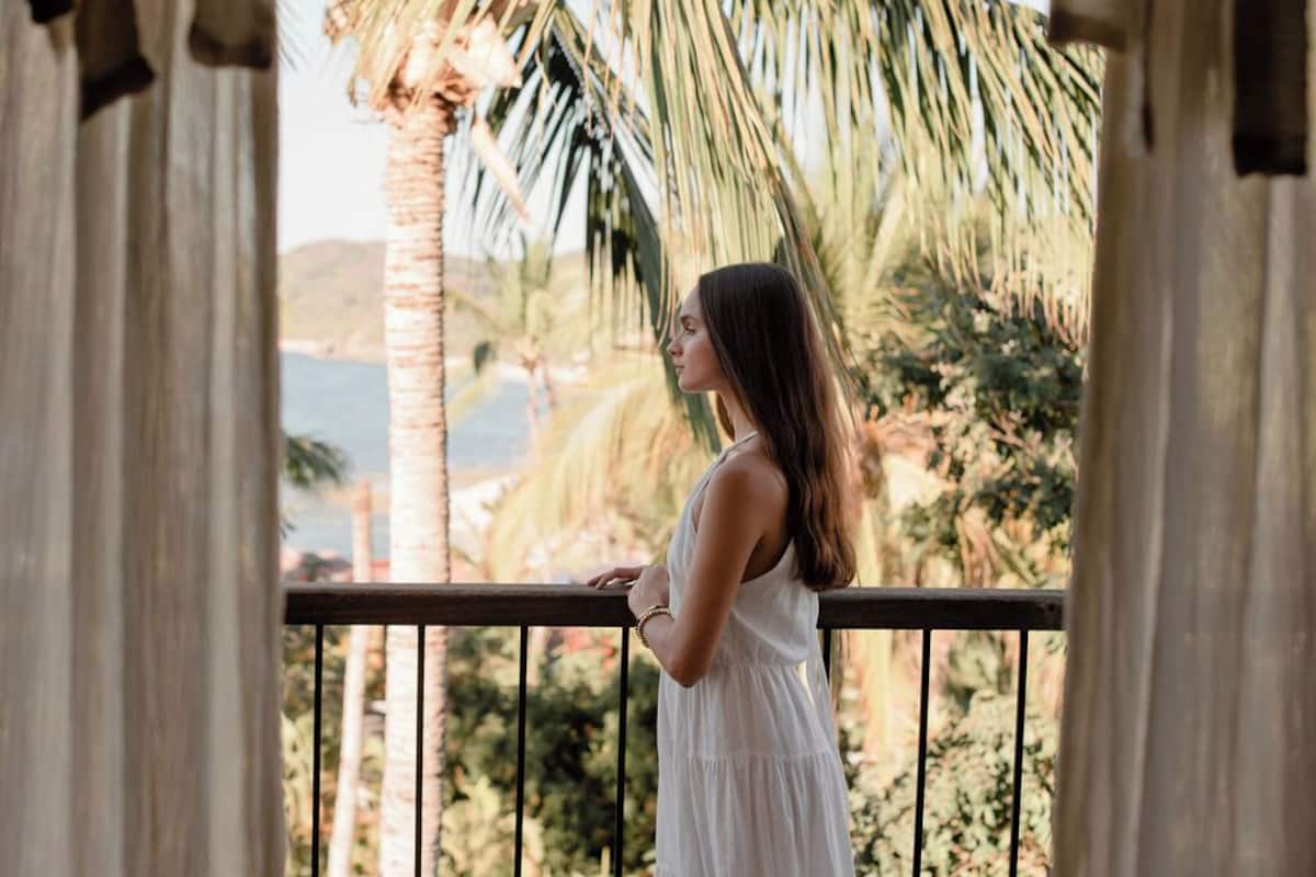 A person standing on a balcony with a view of palm trees and a body of water.