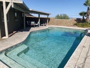 Ein Schwimmbecken mit klarem Wasser, umgeben von Steinmauern und einer überdachten Terrasse mit Stühlen.