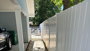 A pathway with stone slabs and a white fence, leading to a garage with a car and a green trash bin. - BSL Living - Across from Pickleball Courts, Splash Pad and Park - Permit #BSL140 (Bay St Louis)