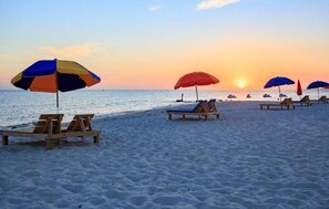 Une plage avec des chaises longues en bois et des parasols, un coucher de soleil en arrière-plan et une mer calme.