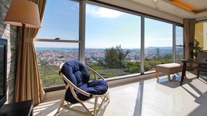 Amplia habitación con grandes ventanales que ofrecen vistas a la ciudad, un sillón cómodo, una mesa y una lámpara.