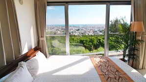 Habitación de hotel con un ventanal que ofrece vistas a la ciudad, una cama con ropa blanca y una planta en maceta.