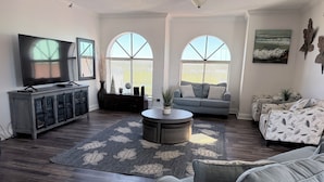 Ein Wohnzimmer mit Fernseher, Sofa und Couchtisch. Zwei Fenster mit Jalousien, eine Pflanze und ein Bild an der Wand sind ebenfalls vorhanden.