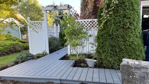 Eine Holzterrasse mit weißem Gitterzaun, einer Pergola und einem gepflegten Garten mit verschiedenen Pflanzen und einem Steinweg.
