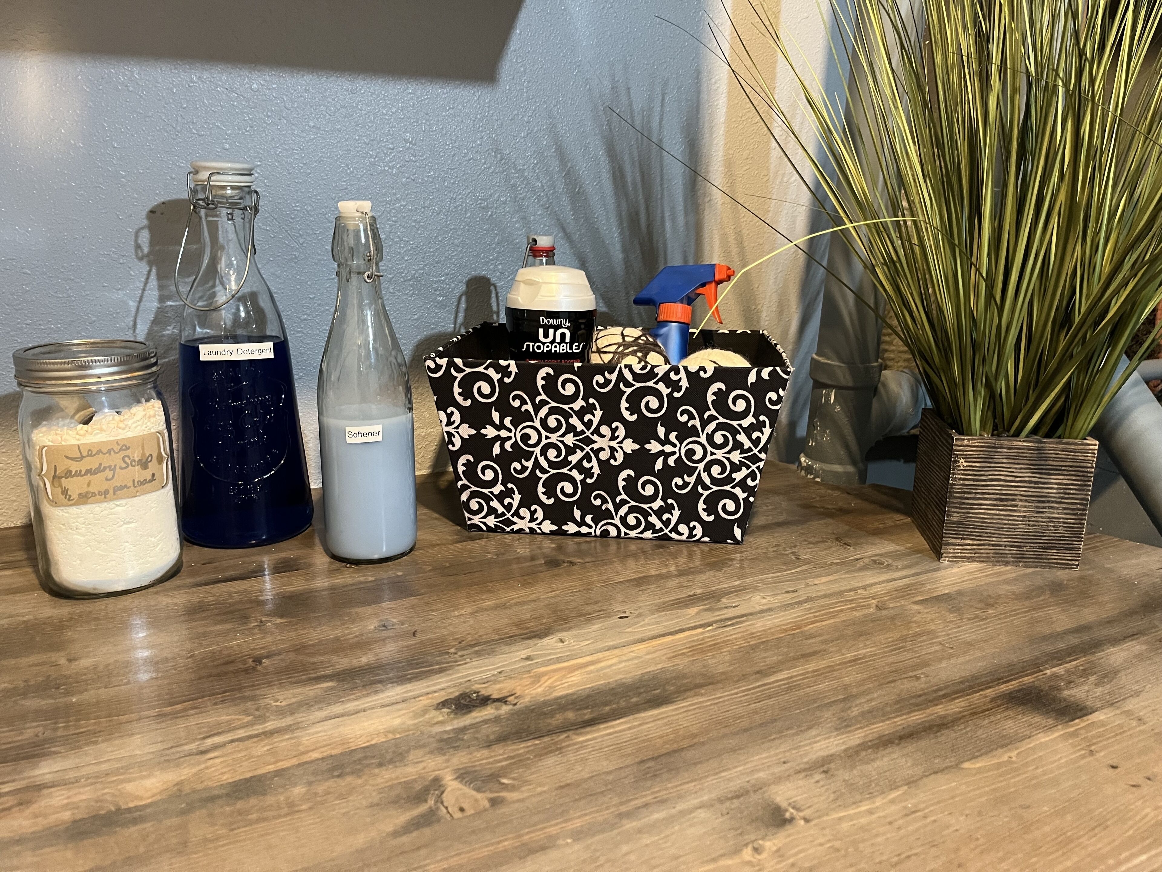 Une surface en bois sur laquelle se trouvent un bocal en verre étiqueté « Savon à lessive », une bouteille étiquetée « Détergent à lessive », une bouteille étiquetée « Adoucissant » et un sac à motifs noir et blanc contenant un flacon pulvérisateur et un récipient.