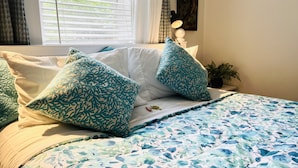 Ein ordentlich bezogenes Bett mit einem blumigen Überwurf und passenden Kissen, ein Fenster mit Jalousien und ein Gemälde an der Wand.