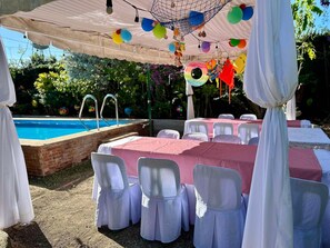 An outdoor pool area with tables and chairs, surrounded by greenery and a clear blue sky.