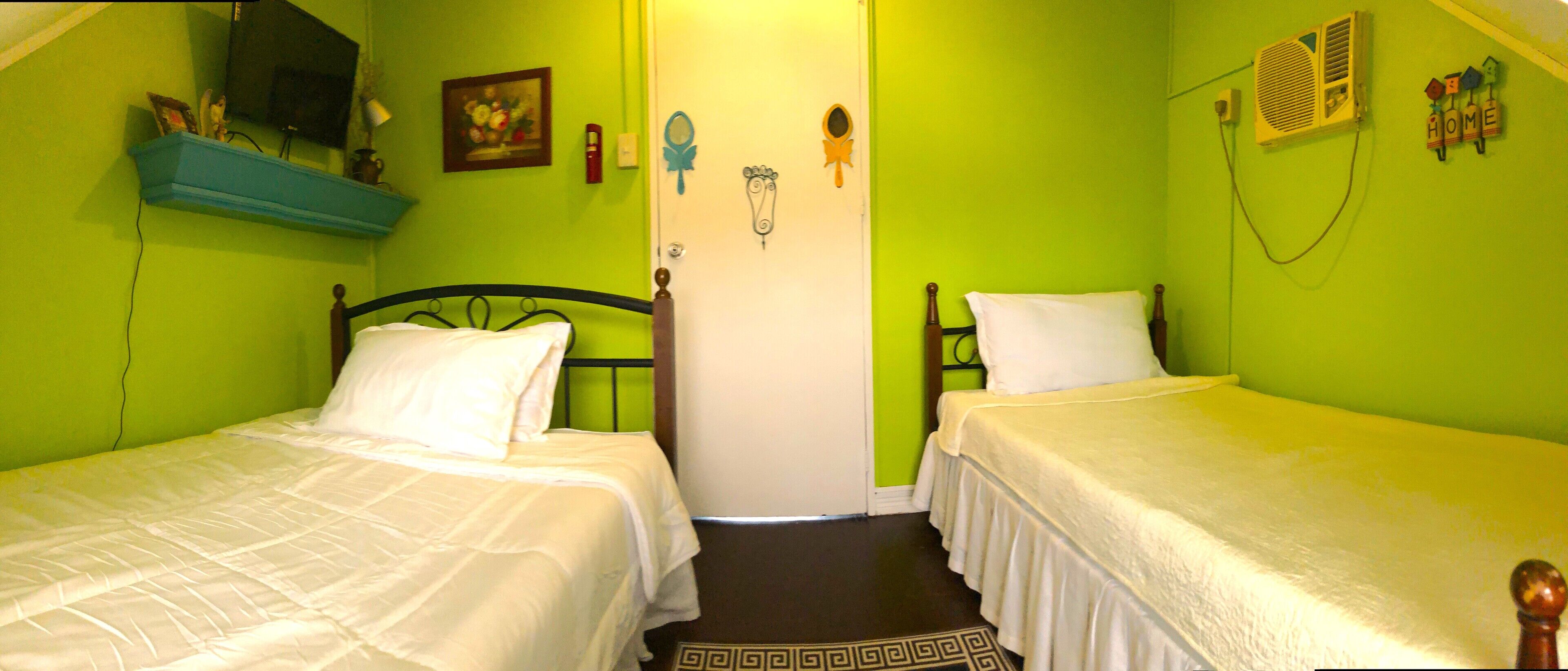 A room with two single beds, a small shelf with books and a vase, a painting on the wall, and a door with a mirror and decorative items.